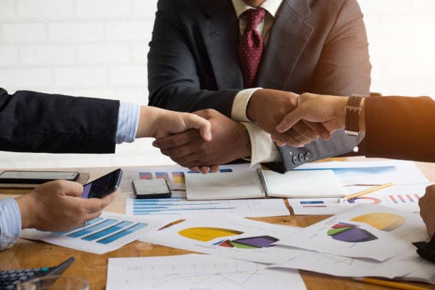 Business partners shaking hands over a table of charts and reports